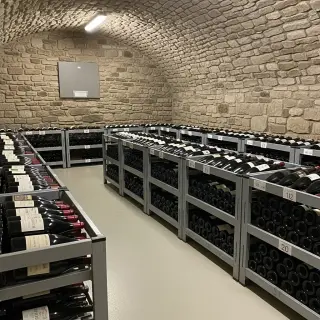 Casiers métalliques de stockage à vin numérotés dans une cave mutualisée avec bouteilles alignées, mur en pierres en arrière-plan et panneau d’informations à gauche.