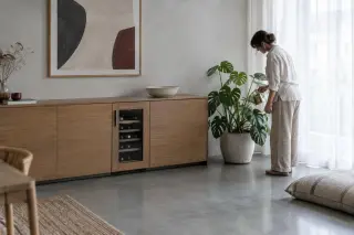Dans un appartement design au béton ciré, une femme arrose une plante posée près d'une cave à vin intégrée comme un meuble bas
