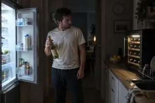 Un homme hésite le soir entre la porte ouverte de son frigo et une petite cave à vin, une bouteille de blanc à la main