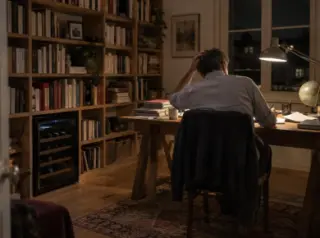 Un homme travaille de dos à son bureau le soir, une mini cave à vin sous la bibliothèque, lampe d'architecte allumée
