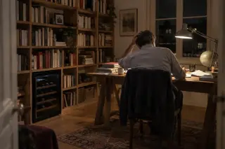 Un homme travaille de dos à son bureau le soir, une mini cave à vin sous la bibliothèque, lampe d'architecte allumée