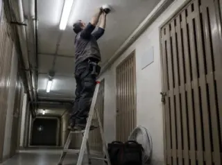 Vu d'en bas, un installateur sur une échelle fixe une caméra au plafond du couloir des caves d'un immeuble