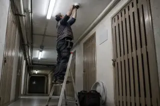 Vu d'en bas, un installateur sur une échelle fixe une caméra au plafond du couloir des caves d'un immeuble