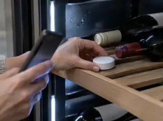 Gros plan sur un petit capteur d'humidité connecté posé sur une clayette de cave à vin, une femme regarde son téléphone