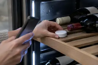 Gros plan sur un petit capteur d'humidité connecté posé sur une clayette de cave à vin, une femme regarde son téléphone