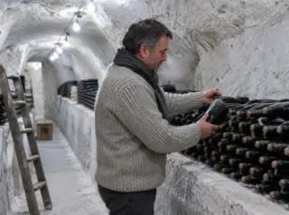 Dans une cave creusée dans la craie, un homme en pull épais prend une bouteille de champagne parmi celles couchées