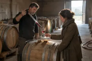 Dans un chai, un vigneron sert à la pipette un vin rouge tiré d'un fût à une visiteuse qui tend son verre