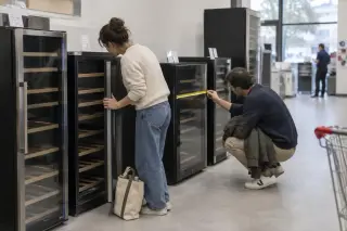 Un couple compare des caves à vin dans un magasin d'électroménager, l'une ouvre une porte vitrée, l'autre mesure la largeur au mètre ruban