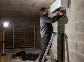 Un installateur fixe au mur d'une cave en parpaing l'unité intérieure d'un climatiseur de cave, perceuse et lampe de chantier