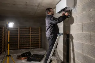 Un installateur fixe au mur d'une cave en parpaing l'unité intérieure d'un climatiseur de cave, perceuse et lampe de chantier
