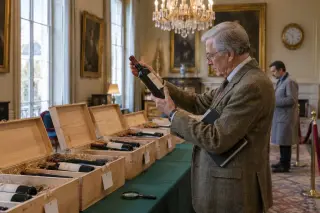 Dans une salle des ventes, un homme âgé examine une bouteille devant des caisses de douze en bois alignées sous un lustre