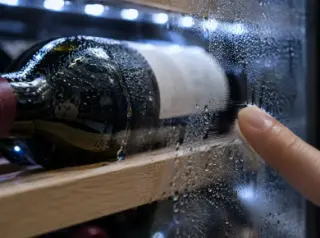 Gros plan sur des gouttes de condensation sur une bouteille et la vitre intérieure d'une cave, un doigt les essuie
