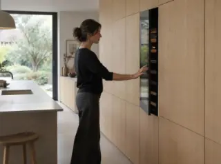 Dans une cuisine d'architecte sans poignées, une femme ouvre par pression une cave à vin affleurante dans le mur de placards