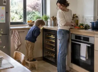 Dans une cuisine familiale à midi, un enfant sur un tabouret regarde les bouteilles d'une cave à vin sous plan à côté du four, sa mère cuisine