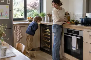 Dans une cuisine familiale à midi, un enfant sur un tabouret regarde les bouteilles d'une cave à vin sous plan à côté du four, sa mère cuisine