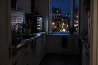 Une cuisine étroite la nuit, la cave à vin allumée, la fenêtre ouverte sur les lumières de la ville