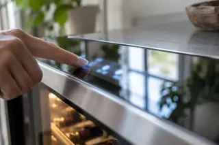 Gros plan sur une main qui touche l'écran tactile flou d'une cave à vin connectée, reflet de la pièce