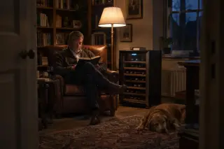 Un homme lit le soir dans un fauteuil, une lampe allumée, une cave à vin posée à côté du fauteuil dans son bureau