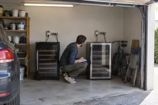 Dans un garage, un homme compare deux caves à vin côte à côte branchées chacune sur un wattmètre