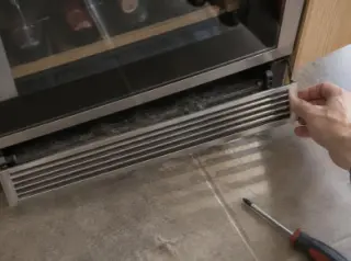 Gros plan vu de dessus sur une main qui retire la grille de ventilation en plinthe sous une cave à vin encastrée