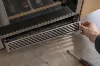 Gros plan vu de dessus sur une main qui retire la grille de ventilation en plinthe sous une cave à vin encastrée