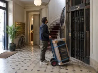 Un homme attend l'ascenseur d'un immeuble haussmannien avec une cave à vin sanglée sur un diable, boîtes aux lettres