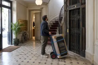 Un homme attend l'ascenseur d'un immeuble haussmannien avec une cave à vin sanglée sur un diable, boîtes aux lettres