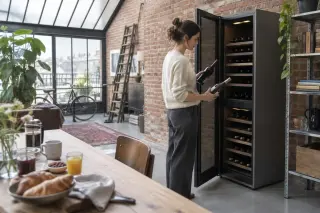 Dans un loft à verrière et briques, une femme sort deux bouteilles d'une cave à vin à deux zones, le matin