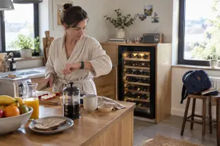 Une femme regarde sa montre en préparant le petit-déjeuner dans sa cuisine ouverte, une cave à vin derrière elle