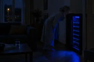 La nuit, une femme en robe de chambre penche l'oreille vers sa cave à vin dont seule la LED bleue éclaire le salon