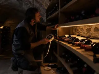 Un électricien colle un ruban LED sous une étagère de cave, les bouteilles éclairées à mi-hauteur, le reste dans la pénombre