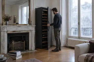 Un homme ouvre une grande cave à vin dans un salon haussmannien au parquet en point de Hongrie, cheminée de marbre, matin d'hiver