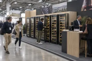Dans une halle d'exposition, des armoires de vieillissement alignées sur un stand, des visiteurs à badges passent