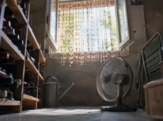 Vu d'en bas, un soupirail de cave ouvert avec un rideau de perles, un petit ventilateur posé au sol, contre-jour d'été