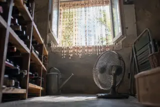 Vu d'en bas, un soupirail de cave ouvert avec un rideau de perles, un petit ventilateur posé au sol, contre-jour d'été