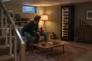 Un homme s'assied sur un canapé dans un sous-sol aménagé, une cave à vin colonne sous une lampe, tapis