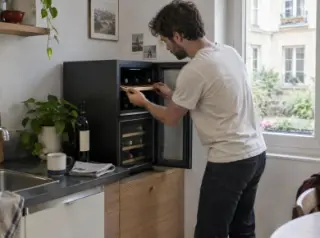 Dans la kitchenette d'un studio, un homme ajuste une clayette d'une cave à vin compacte à plusieurs températures posée sur un meuble bas