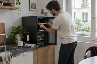 Dans la kitchenette d'un studio, un homme ajuste une clayette d'une cave à vin compacte à plusieurs températures posée sur un meuble bas