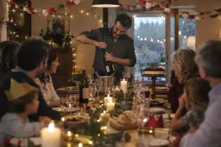 Un homme debout débouche une bouteille au bout d'une grande table de fête décorée de guirlandes, convives flous