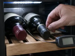 Gros plan sur une sonde de thermomètre glissée entre deux bouteilles dans une cave à vin, afficheur flou, main d'homme