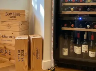 Home wine fridge set about 10 cm from the wall, measuring tape on the floor, in a cozy kitchen corner with wine bottles inside and boxes nearby