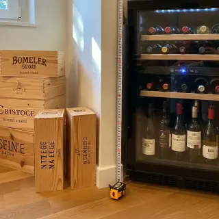 Home wine fridge set about 10 cm from the wall, measuring tape on the floor, in a cozy kitchen corner with wine bottles inside and boxes nearby