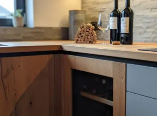 Built-in wine cellar in a modern kitchen surrounded by wood and MDF cabinetry, wine bottles and glass on the countertop, warm natural lighting.