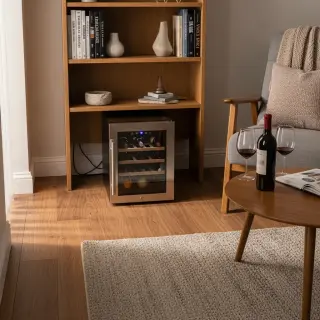 Electric wine fridge in a cozy living room corner on a rug, next to bookshelf and armchair, with bottles and glasses, in natural daylight