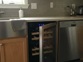 Small refrigerated wine cellar under kitchen counter between sink and dishwasher, glass door open showing bottles on wooden shelves