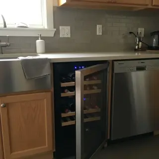 Small refrigerated wine cellar under kitchen counter between sink and dishwasher, glass door open showing bottles on wooden shelves