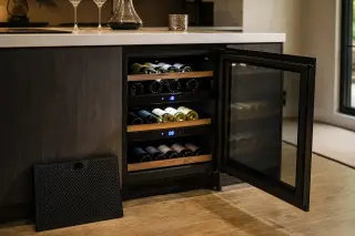 Compact electric multi-zone wine cellar under a modern kitchen counter, open to show organized bottles in different temperature zones, with digital controls and soft interior lighting.