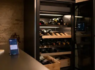 Electric wine cellar in a home setting with bottles on wooden racks, glass door and touchscreen display, smartphone showing cellar app in foreground.