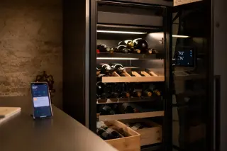 Electric wine cellar in a home setting with bottles on wooden racks, glass door and touchscreen display, smartphone showing cellar app in foreground.