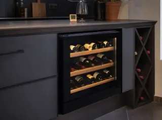 Compact electric wine cellar under a kitchen counter with wine bottles neatly organised, anti-UV glass door, digital hygrometer nearby, and a glass of red wine on the countertop in a modern urban kitchen.